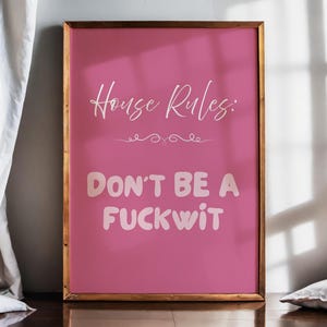 May include: A framed pink poster with the text "House Rules: DON'T BE A FUCKWIT" in white. The poster has a gold-colored frame and is leaning against a wall. A white curtain and pillow are visible.