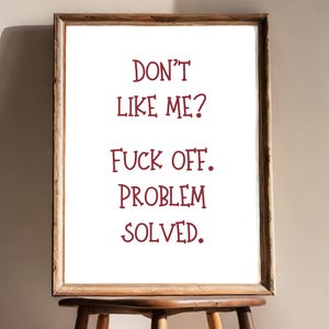May include: A framed print with the text "DON'T LIKE ME? FUCK OFF. PROBLEM SOLVED." in burgundy lettering. The wooden frame is a warm brown, and the print is displayed on a wooden stool.