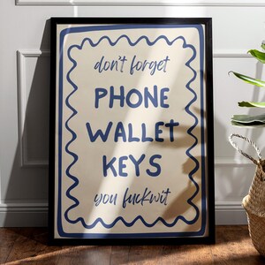 May include: A framed poster with a cream-colored background and blue text that reads "don't forget PHONE WALLET KEYS you fuckwit." The text is surrounded by a blue wavy border. A plant in a woven basket is in the background.