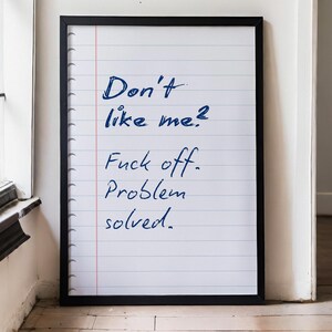 May include: A framed print featuring a white background with horizontal blue lines and a vertical red margin line, resembling a sheet of notebook paper. The text "Don't like me? Fuck off. Problem solved." is written in dark blue, handwritten-style script.