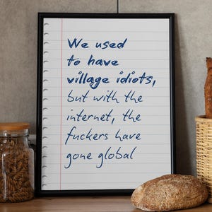 May include: A black framed wall art piece featuring a white background with blue handwritten text that reads: We used to have village idiots, but with the internet, the fuckers have gone global. The paper design mimics a lined notebook page.
