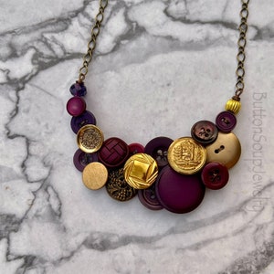 May include: A purple and gold button necklace with a variety of different buttons, including some with intricate designs. The necklace is made with a brass chain and has a purple bead at the top.