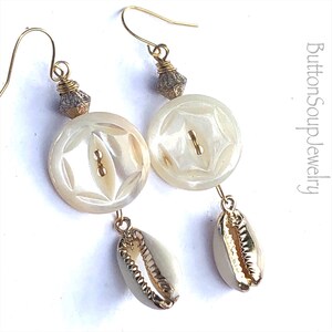 May include: A pair of gold earrings with white shell buttons and cowrie shells. The buttons have a carved design and the cowrie shells have a gold trim.