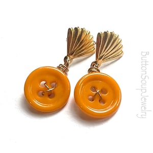 May include: A pair of orange button earrings with gold fan-shaped accents. The earrings are made with vintage buttons and are a unique and stylish accessory.
