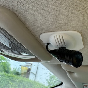 Può includere: Un microfono nero con corpo cilindrico è montato su una base di plastica bianca, fissata al soffitto del veicolo. Il microfono è collegato tramite un filo nero. È visibile anche un'apparecchio di illuminazione.