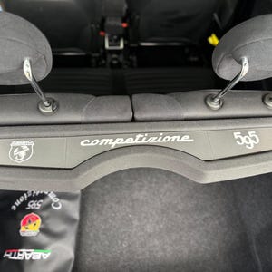 Può includere: Interno auto, vista dei poggiatesta posteriori e un pannello grigio con la scritta "competizione" in bianco. Il pannello mostra anche "595", una striscia tricolore e il logo Abarth. Lo schema dei colori è grigio scuro.