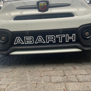 Può includere: Un'auto bianca con un logo "ABARTH" bianco e nero sulla griglia anteriore. L'auto ha un logo "ABARTH" nero e rosso sul cofano.