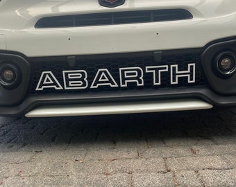 Può includere: Un'auto bianca con un logo "ABARTH" bianco e nero sulla griglia anteriore. L'auto ha un logo "ABARTH" nero e rosso sul cofano.