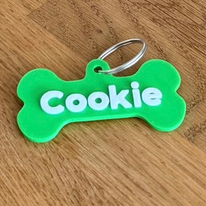 Può includere: Una targhetta per animali domestici a forma di osso verde brillante con il nome Cookie in lettere bianche in rilievo. La targhetta include un anello metallico argentato fissato all'occhiello superiore per agganciarla a un collare.