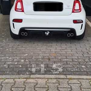 Könnte beinhalten: Ein weißer Fiat 500 Abarth mit schwarzem Heckstoßfänger und zwei Auspuffrohren. Das Auto hat einen schwarzen Heckspoiler und einen schwarzen Heckdiffusor. Das Auto steht auf einer grauen Pflasterfläche.
