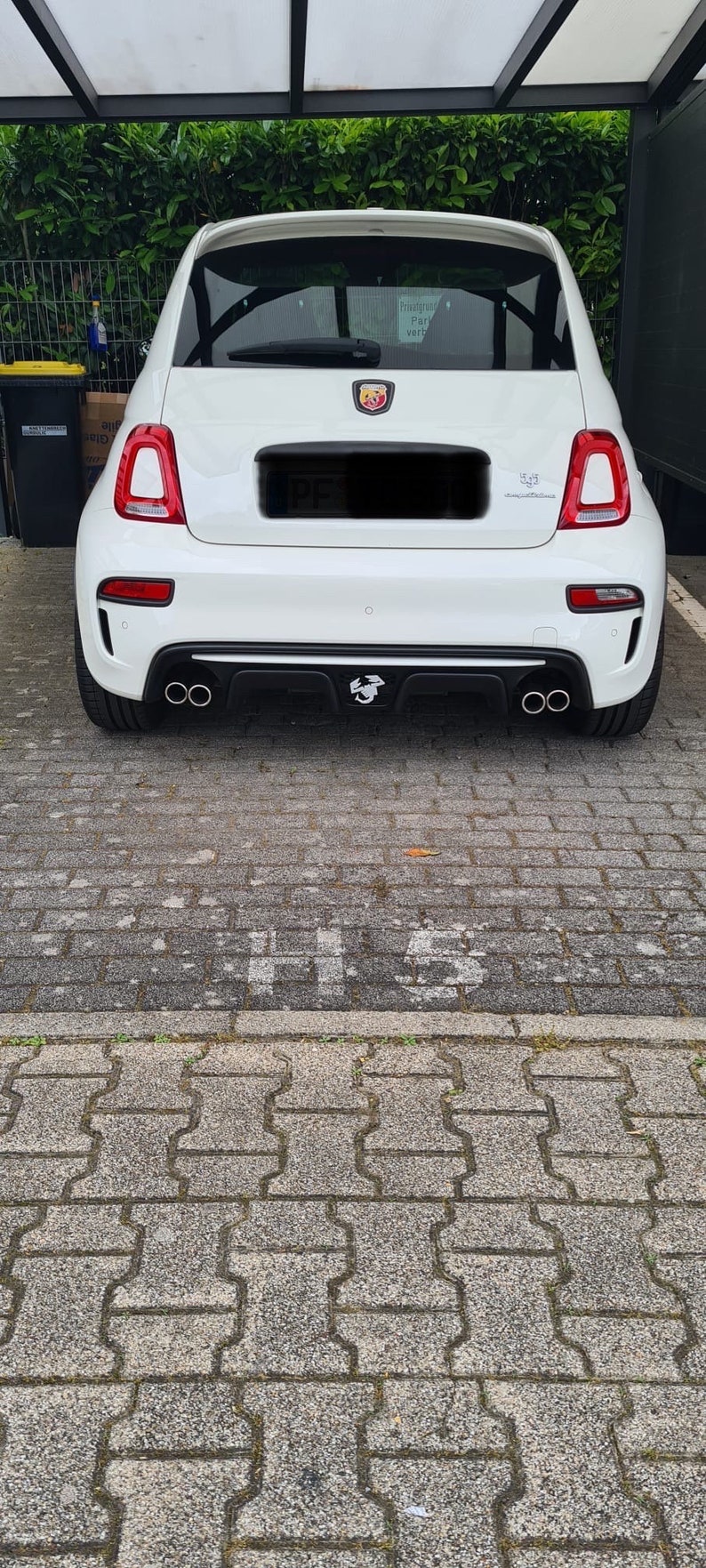 Pu&ograve; includere: Una Fiat 500 Abarth bianca con un paraurti posteriore nero e due scarichi. L'auto ha uno spoiler posteriore nero e un diffusore posteriore nero. L'auto &egrave; parcheggiata su un vialetto di mattoni grigi.