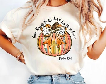 May include: A white t-shirt with a graphic of a pumpkin with a bow and a cross. The text "Give Thanks To The Lord For He Is Good" is written around the pumpkin. The text "Psalm 136:1" is written below the pumpkin.