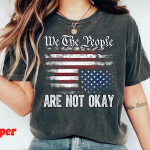 May include: A dark gray t-shirt with a distressed look featuring a faded American flag and the text "We the People Are Not Okay".