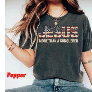 May include: A dark gray t-shirt with a distressed look featuring the text "JESUS MORE THAN A CONQUEROR" in white letters with a red, white, and blue American flag design behind the text.