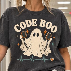 May include: A charcoal gray short-sleeve t-shirt featuring a graphic of a white ghost holding two defibrillator paddles. The text "CODE BOO" is printed above the ghost in a cream font, with a teal heartbeat line and a small orange heart below.