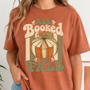 May include: A rust-colored short-sleeve t-shirt featuring a graphic of a pumpkin made of stacked books. The text "ALL BOOKED for FALL" is printed in a retro font in shades of sage green and cream, centered above and below the pumpkin illustration.