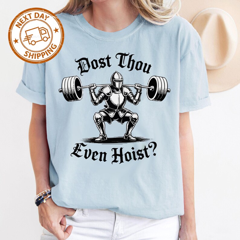 May include: Light blue t-shirt featuring a black and white illustration of a knight lifting a barbell. The text "Dost Thou Even Hoist?" is printed below the illustration. The shirt has rolled-up sleeves.