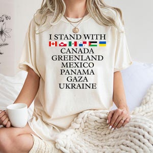 Puede incluir: Una camiseta blanca con el texto "I stand with" y una fila de banderas, seguida del texto "Canad&aacute;, Groenlandia, M&eacute;xico, Panam&aacute;, Gaza, Ucrania".