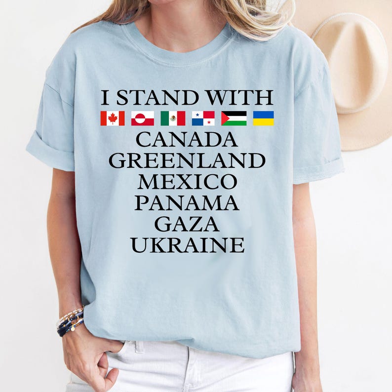 Puede incluir: Camiseta azul claro con el texto "I Stand With" y una fila de banderas sobre el texto. Debajo de las banderas, el texto dice "Canad&aacute;, Groenlandia, M&eacute;xico, Panam&aacute;, Gaza, Ucrania".