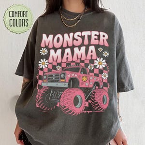 以下が含まれることがあります： モンスタートラックと花、そして「モンスターママ」という文字がピンクで描かれたダークグレーのTシャツです。