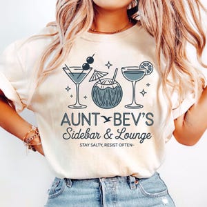 May include: Cream-colored t-shirt featuring a cocktail-themed design. The shirt displays illustrations of a martini, a coconut drink, and a cocktail with a lemon slice. The text "AUNT BEV'S Sidebar & Lounge" is printed below the illustrations, with the phrase "STAY SALTY, RESIST OFTEN-" below.