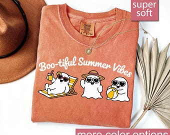 May include: Coral-colored t-shirt with the phrase "Boo-tiful Summer Vibes" and illustrations of cartoon ghosts enjoying summer. The shirt has a crew neck and short sleeves. A gold necklace is draped on the shirt. The shirt is labeled "super soft".
