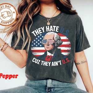 May include: Dark gray t-shirt featuring a graphic of George Washington wearing American flag sunglasses, set against a backdrop of the American flag. The text reads "THEY HATE U.S. CUZ THEY AIN'T U.S."