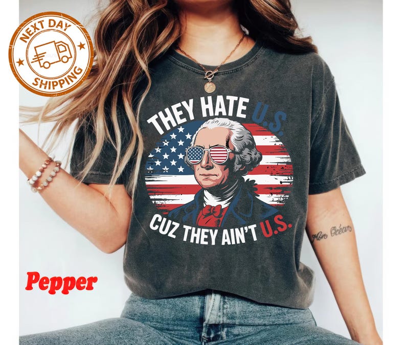 May include: Dark gray t-shirt featuring a graphic of George Washington wearing American flag sunglasses, set against a backdrop of the American flag. The text reads "THEY HATE U.S. CUZ THEY AIN'T U.S."