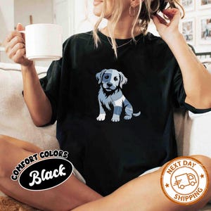 May include: Black t-shirt featuring a blue and white graphic of a puppy. The puppy design has a patchwork pattern. The shirt has the words "COMFORT COLORS Black" in a circular design. A "NEXT DAY SHIPPING" logo is also visible.