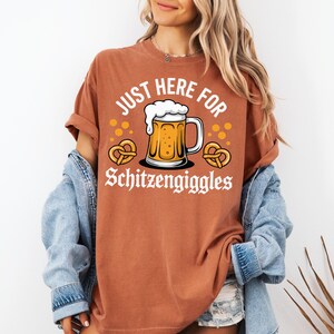 Peut inclure: T-shirt rouille avec l'inscription "JUST HERE FOR Schitzengiggles" et un dessin de chope de bi&egrave;re et de bretzels. Le t-shirt a des manches retrouss&eacute;es et est port&eacute; avec une veste en jean.