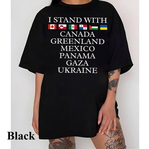 Puede incluir: Camiseta negra de gran tama&ntilde;o con el texto "I Stand With" y una fila de banderas que representan a Canad&aacute;, Groenlandia, M&eacute;xico, Panam&aacute;, Gaza y Ucrania.