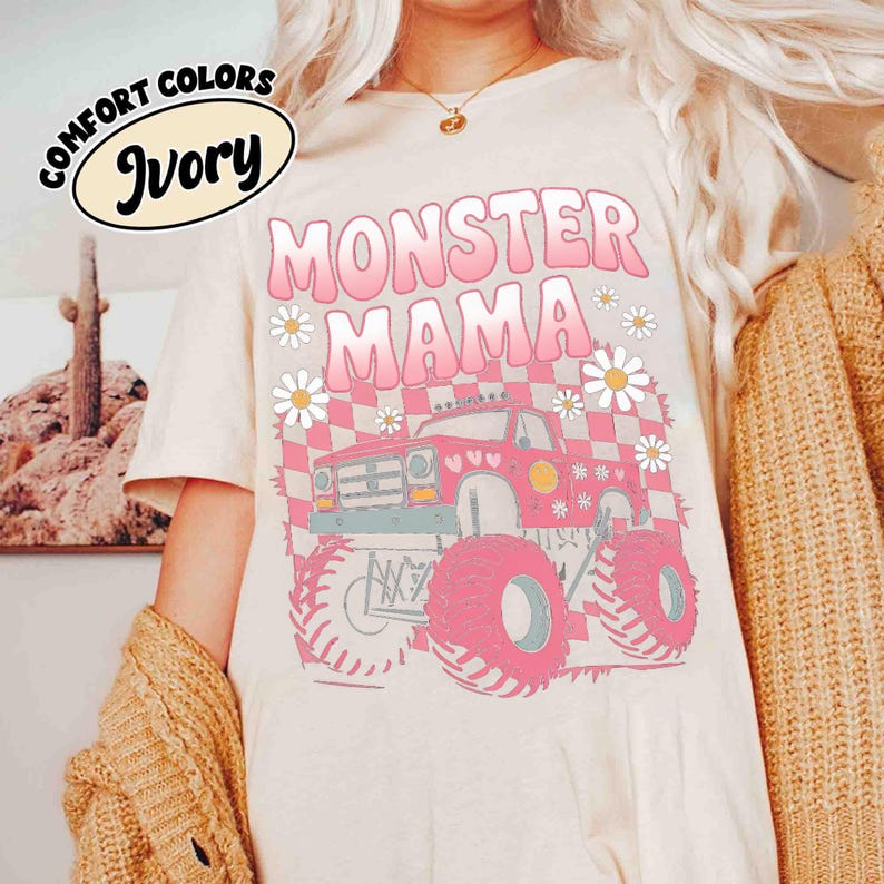 以下が含まれることがあります： モンスタートラックのピンクと白のグラフィックが描かれた白いTシャツ。トラックの上には「モンスターママ」の文字が書かれています。トラックはピンクの車輪を備え、ピンクと白の市松模様の背景にピンクの花が咲いています。