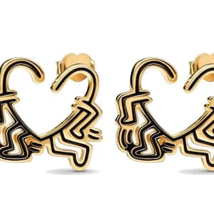 May include: A pair of gold-tone earrings featuring a heart-shaped design with a black outline. The heart is formed by a stylized figure with arms and legs.