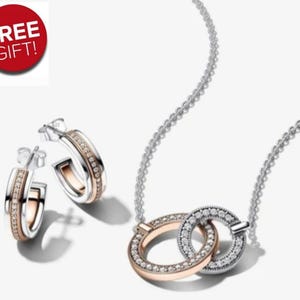 May include: A jewellery set comprising a necklace and earrings. The necklace features two interlocking rings, one rose gold and one silver, both set with small, sparkling stones. The earrings are hoop-style, with a similar design of rose gold and silver with stones.