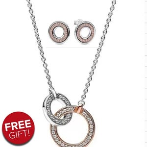 May include: A silver necklace featuring two intertwined rings, one silver and one rose gold, both set with clear crystals. The image also shows matching stud earrings. A red circle with the text "FREE GIFT!" is visible.