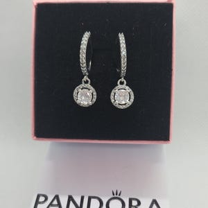 May include: Silver hoop earrings, each featuring a central, square-cut, clear stone encircled by a halo of smaller, clear stones. The earrings are presented in a black box with a pink interior. The brand name "PANDORA" is visible at the bottom.