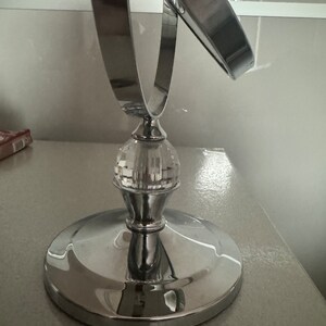 Könnte beinhalten: Ein verchromter Schminkspiegel mit einem Kristallfuß. Der Spiegel ist rund und in einem Winkel geneigt. Der Fuß ist aus klarem Kristall mit einem facettierten Design.