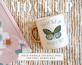 May include: White ceramic mug with a gold handle. The mug has a butterfly design with the word "marché" printed on it. The mug is sitting on a beige knitted blanket.