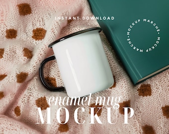 May include: White enamel mug with a black rim and handle, sitting on a pink and brown knitted blanket. The text "enamel mug MOCKUP" is printed below the mug.