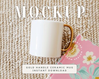 May include: White ceramic mug with a gold handle, sitting on a cream-colored rug next to a pink and white floral notebook. The notebook has the text "MARCHÉ - MOCKUP" on the cover.