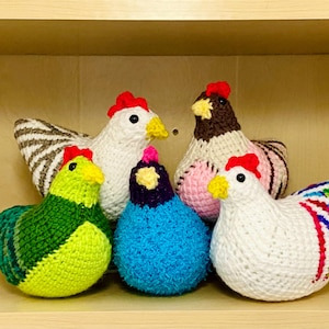 May include: A collection of six handmade crochet chickens in various colors. The chickens have red combs and yellow beaks. The colors include white, green, blue, brown, and pink. The chickens are arranged on a wooden shelf.