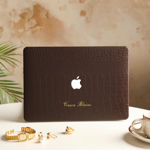 May include: A dark brown laptop case with a textured crocodile skin pattern. The case features a central cutout for an Apple logo and the gold-colored text Vinson Blaine printed below it. The surface has a matte finish and a rectangular shape.