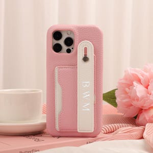 May include: Pink leather phone case with a white card slot and a white strap with the initials "BWM". The case has a textured finish and a camera cutout. A white teacup and saucer are in the background.