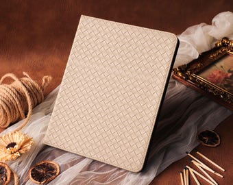 May include: A beige tablet case with a woven pattern. The case is rectangular and has a black border. The image also includes a spool of twine, a dried flower, and a framed picture.