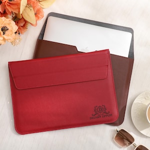 May include: A red leather laptop sleeve with a flap closure, featuring the text "Dreamy Lippert" and a logo. A brown leather sleeve is visible behind it, holding a laptop. A cup of coffee and sunglasses are also on the table.