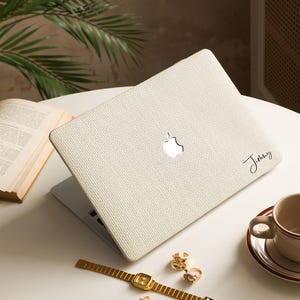 May include: A cream-colored textured laptop skin applied to the lid of a silver laptop. The skin features a visible woven fabric pattern and the name "Jimmy" written in black cursive script on the lower right side, with the Apple logo visible.