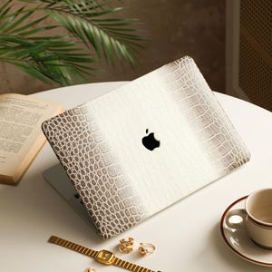 May include: A laptop case featuring a white and beige crocodile skin texture pattern with a gradient effect. The case has a central cutout revealing the black Apple logo. The design includes a textured, embossed surface that mimics reptile scales.