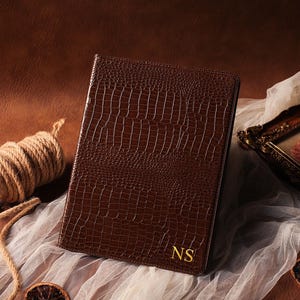 Peut inclure: Un journal en cuir marron avec une texture peau de crocodile. Le journal porte les initiales dorées "NS" dans le coin inférieur droit. Une bobine de ficelle et un tableau encadré sont en arrière-plan.