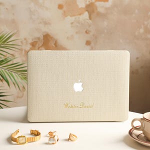 May include: A cream-colored textured laptop case featuring a cutout for the Apple logo and the gold-colored cursive text "Webster Daniel" centered on the front surface. The case has a woven fabric-like appearance with a consistent grid pattern.