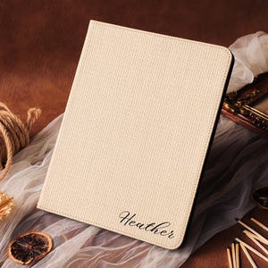 Op de afbeelding: Crèmekleurige tablethoes met de naam "Heather" in cursief schrift. De hoes heeft een getextureerd oppervlak en een zwarte rand. De achtergrond bevat een rol touw, gedroogde bloemen en een ingelijste foto.